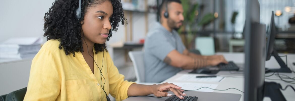A woman in a yellow shirt and a man in grey, both wearing headsets, work at desktop computers in the modern Elite Property London office, focused on delivering exceptional property management and guaranteed rent services.