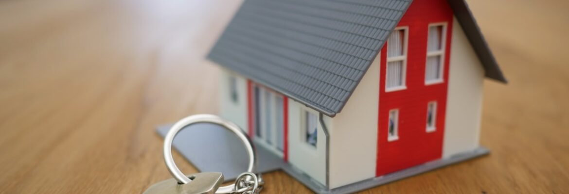 A set of house keys with a keyring lies on a wooden surface next to a small model of a modern house with a red and white exterior.