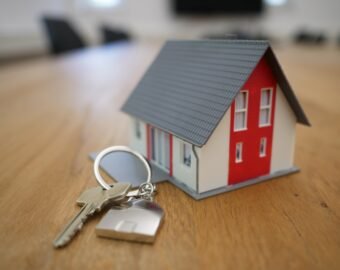 A set of house keys with a keyring lies on a wooden surface next to a small model of a modern house with a red and white exterior.