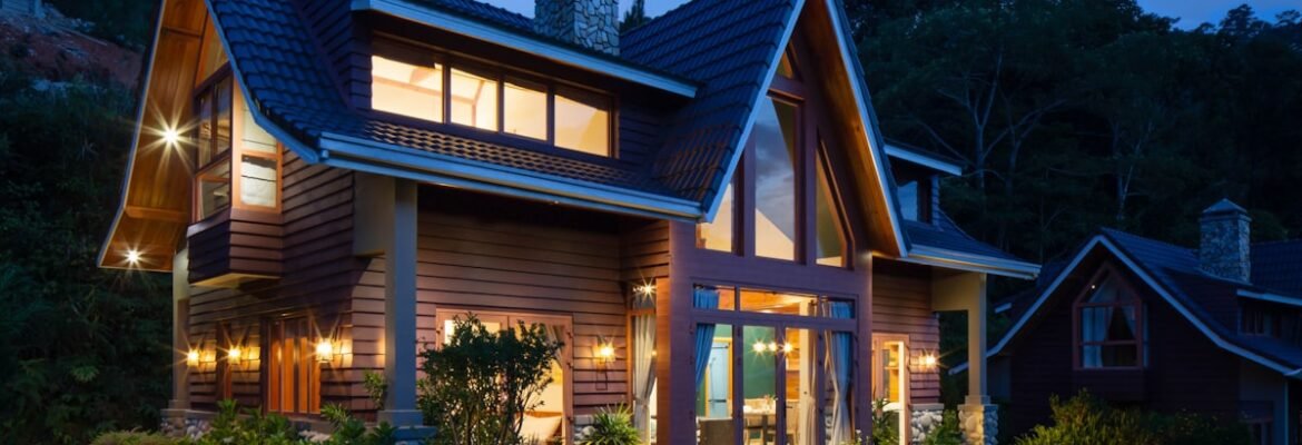 A modern two-storey house with large windows is warmly lit at dusk. The home features timber cladding, a steep roof, and a stone chimney, surrounded by landscaped gardens and greenery.
