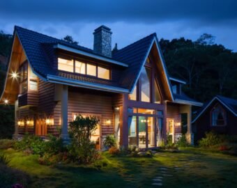 A modern two-storey house with large windows is warmly lit at dusk. The home features timber cladding, a steep roof, and a stone chimney, surrounded by landscaped gardens and greenery.